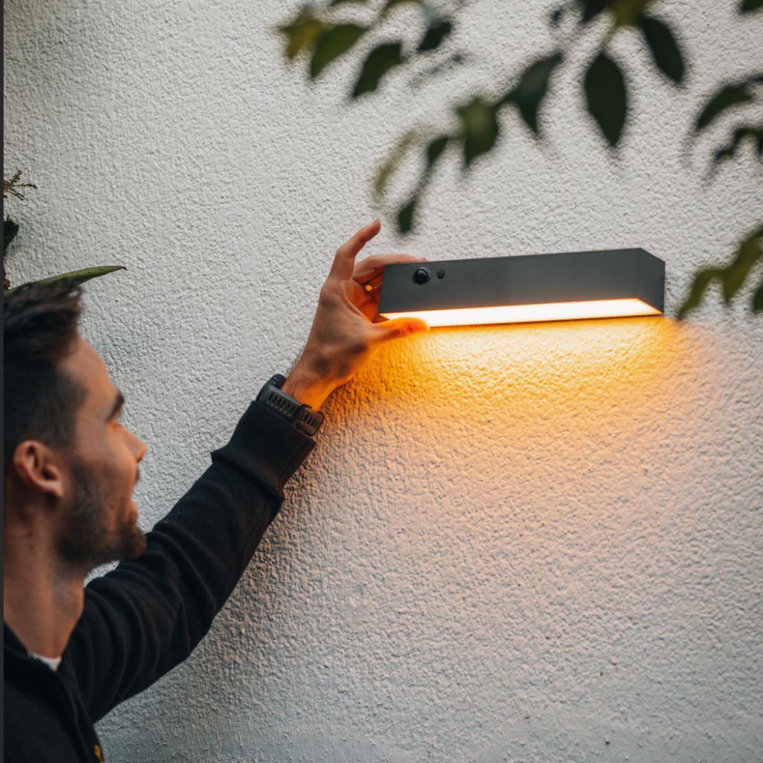 Person adjusting a wall-mounted light fixture on a textured wall with greenery.