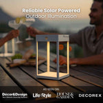 Lanterna solare da esterno con una poltrona a sacco nera e un tavolino di legno su una terrazza in legno; la lanterna diffonde una luce calda. L'immagine include anche dei loghi che indicano che il prodotto è ecologico ed è stato presentato su Décor&Design e Life Style House&Garden.
