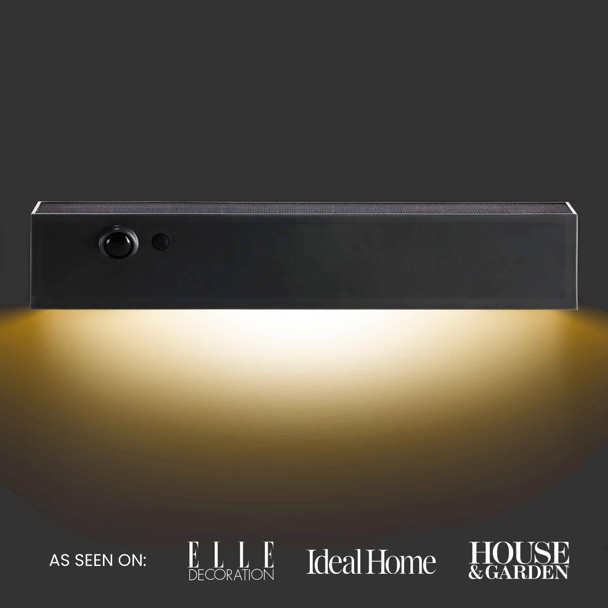 Moderna lampada da parete su sfondo scuro con i loghi di “ELLE Decoration”, “Ideal Home” e “House & Garden”.